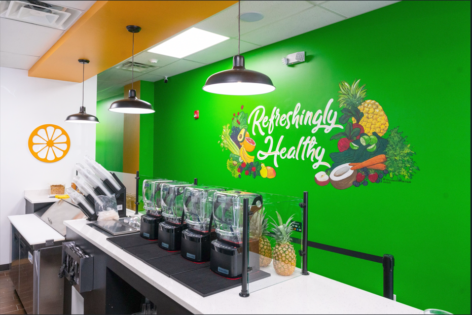 Smoothie bar counter with blenders, hanging lights, and a bright green wall mural reading “Refreshingly Healthy.”