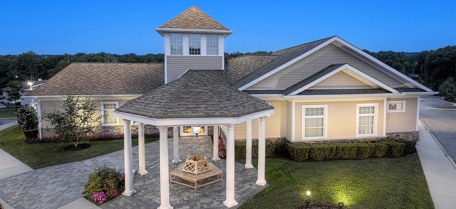 A beige house with a white gazebo, stone patio, and lit landscape at dusk.