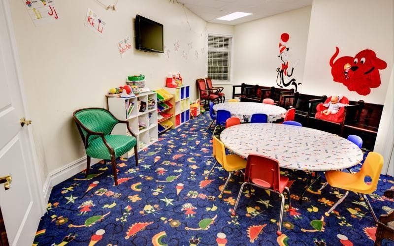 Children’s playroom with colorful carpet, small tables, chairs, toys, and wall art