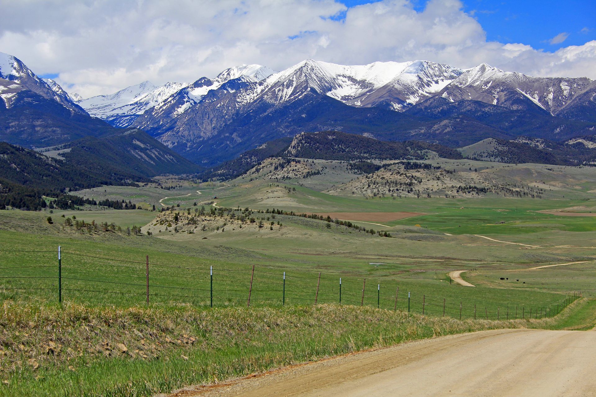 Dirt road leading to green valley with snow-capped mountains and blue sky.
