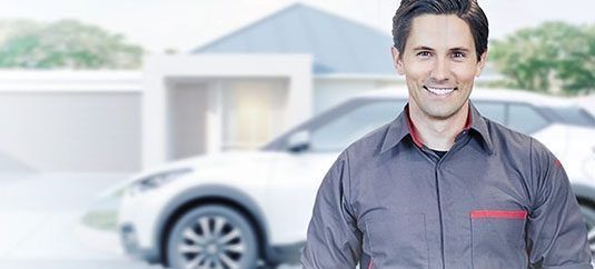 Hombre sonriente con camisa gris de pie junto a un coche plateado en la entrada de una luminosa casa suburbana.