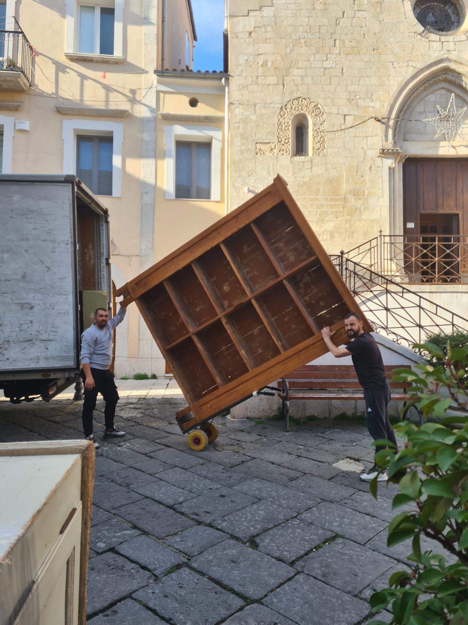 Due uomini inclinano un grande armadio di legno su un carrello con ruote in un cortile di pietra vicino all'ingresso di un edificio.
