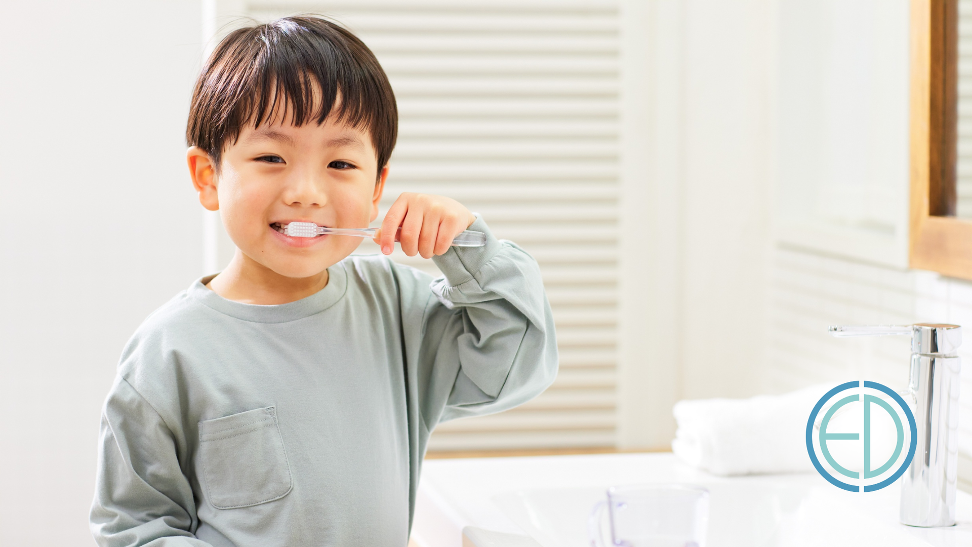 Boy brushing teeth in a bathroom. He's smiling, holding toothbrush. White sink, towel, and mirror visible.