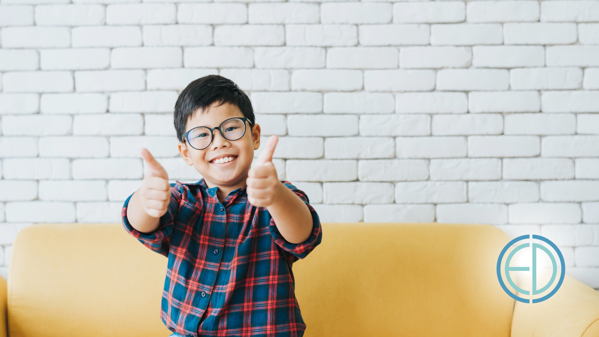 Boy with glasses giving thumbs up, smiling, against a white brick wall and yellow sofa.