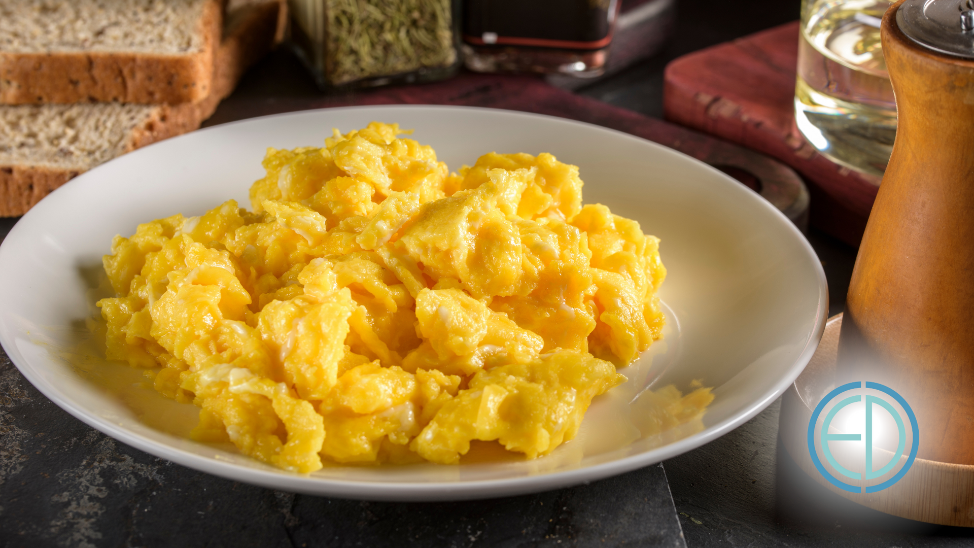 Scrambled eggs on a white plate with slices of bread, oil, and a pepper mill in the background.