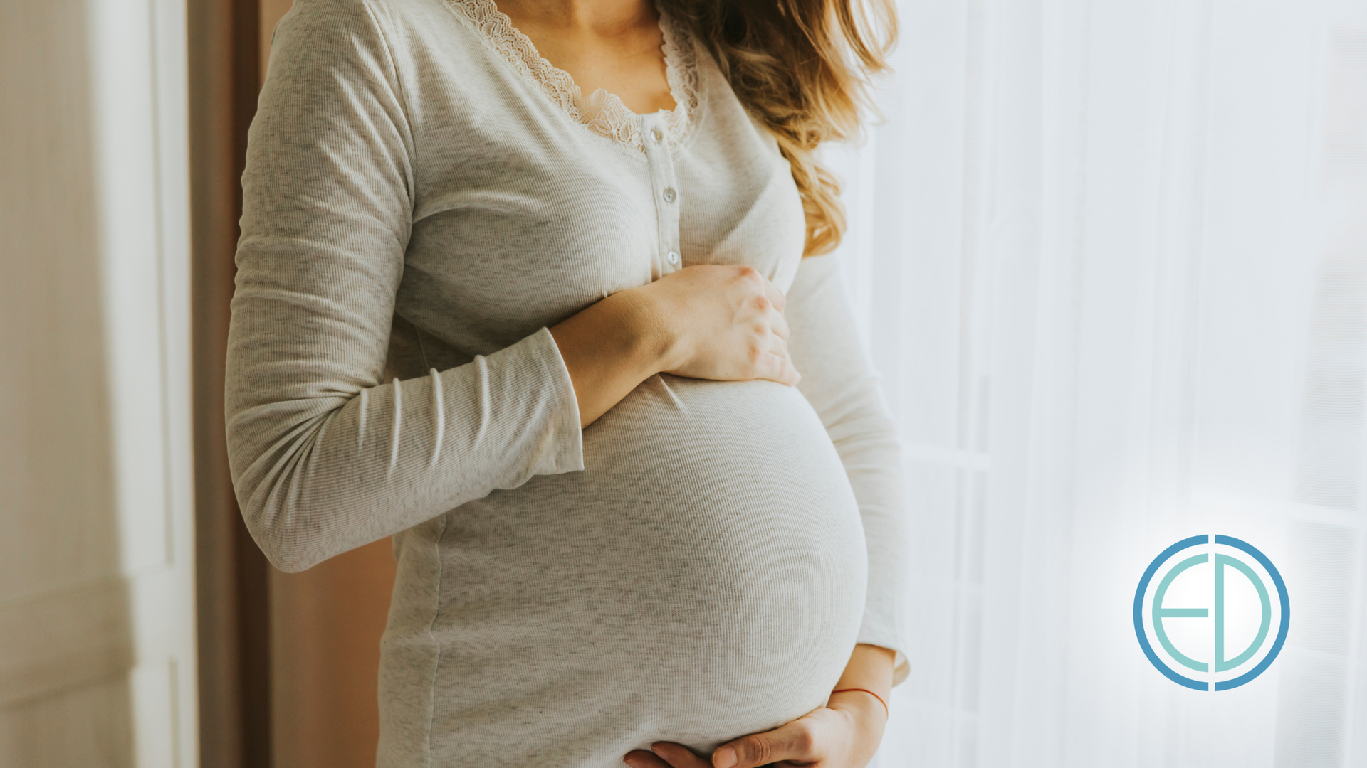 Pregnant person in a gray sweater holding their belly near a bright window