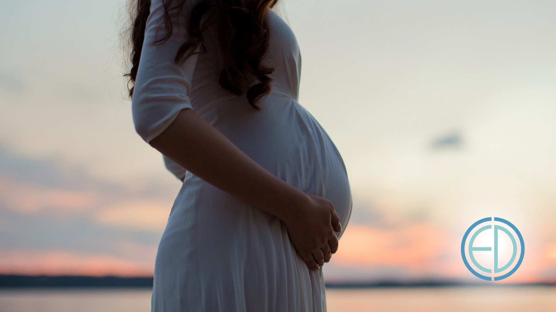 A pregnant woman in a white dress is holding her belly.
