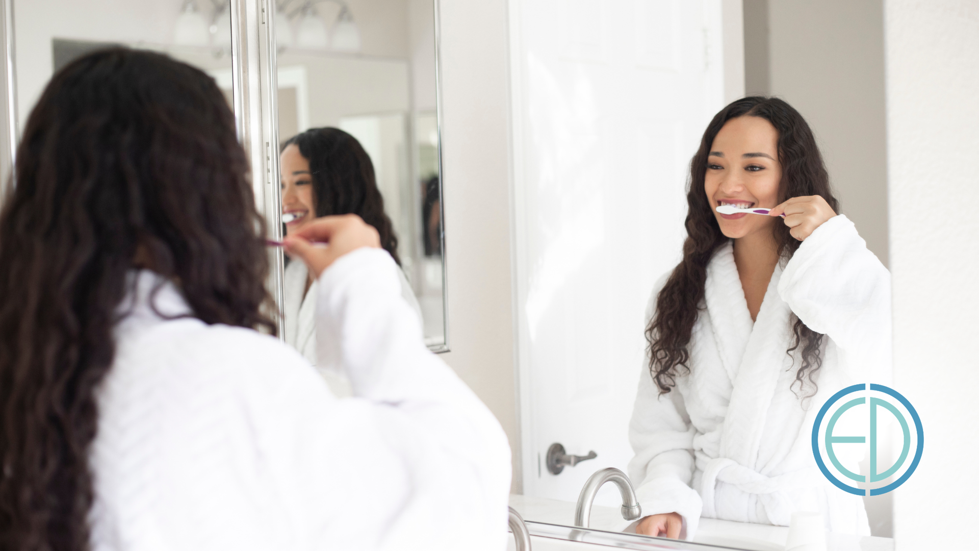 Woman brushing her teeth in a bathroom mirror, wearing a white robe and smiling.