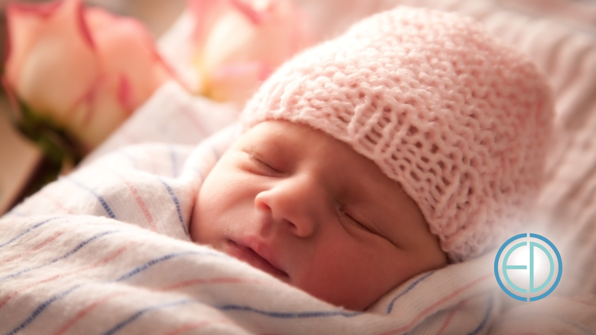 Sleeping newborn wrapped in a striped blanket, wearing a pink knitted hat, with soft flowers in the background