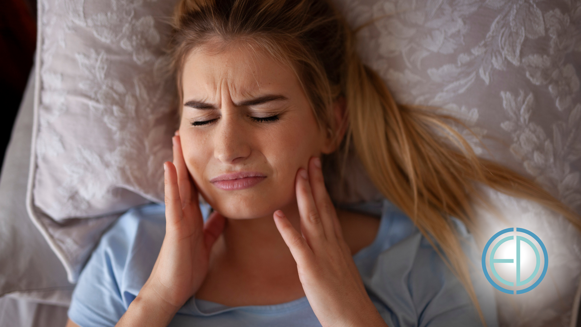 Woman in bed with pained expression, hands on cheeks, indicating possible tooth or jaw pain.