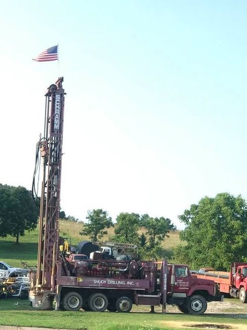 A maroon drilling rig truck with an American flag attached to the top of its extended mast, parked in a grassy field.