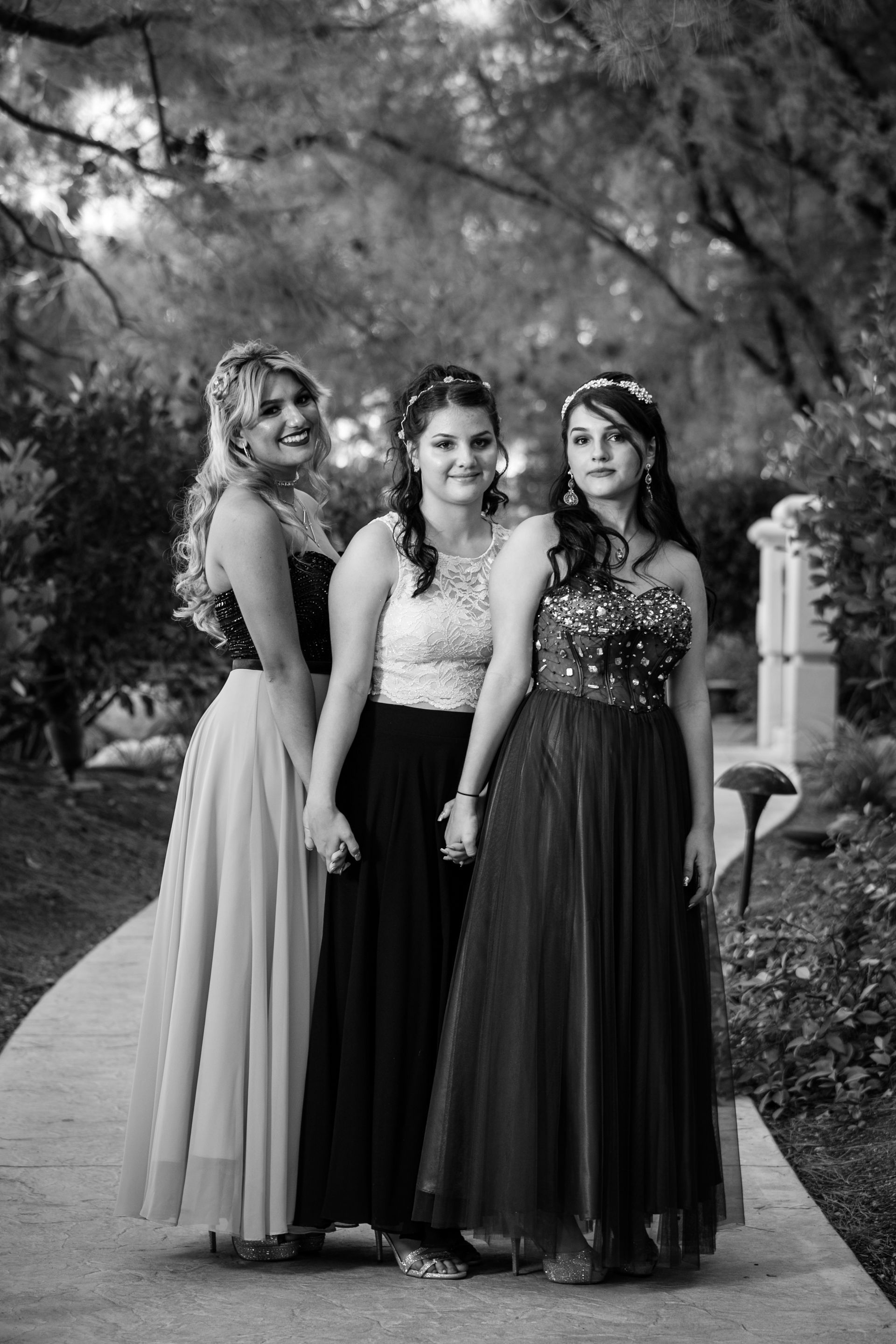 Three women in formal dresses standing on a tree-lined garden path