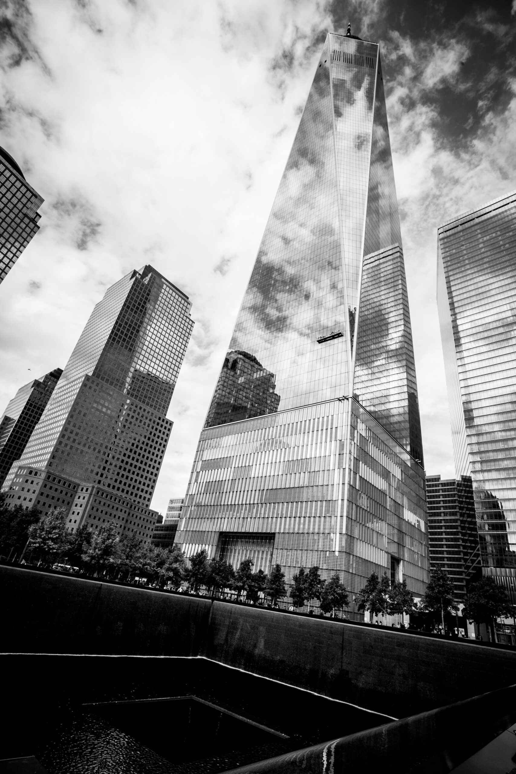 Black-and-white view of skyscrapers and a glass tower in a city plaza under cloudy sky — architectural photography