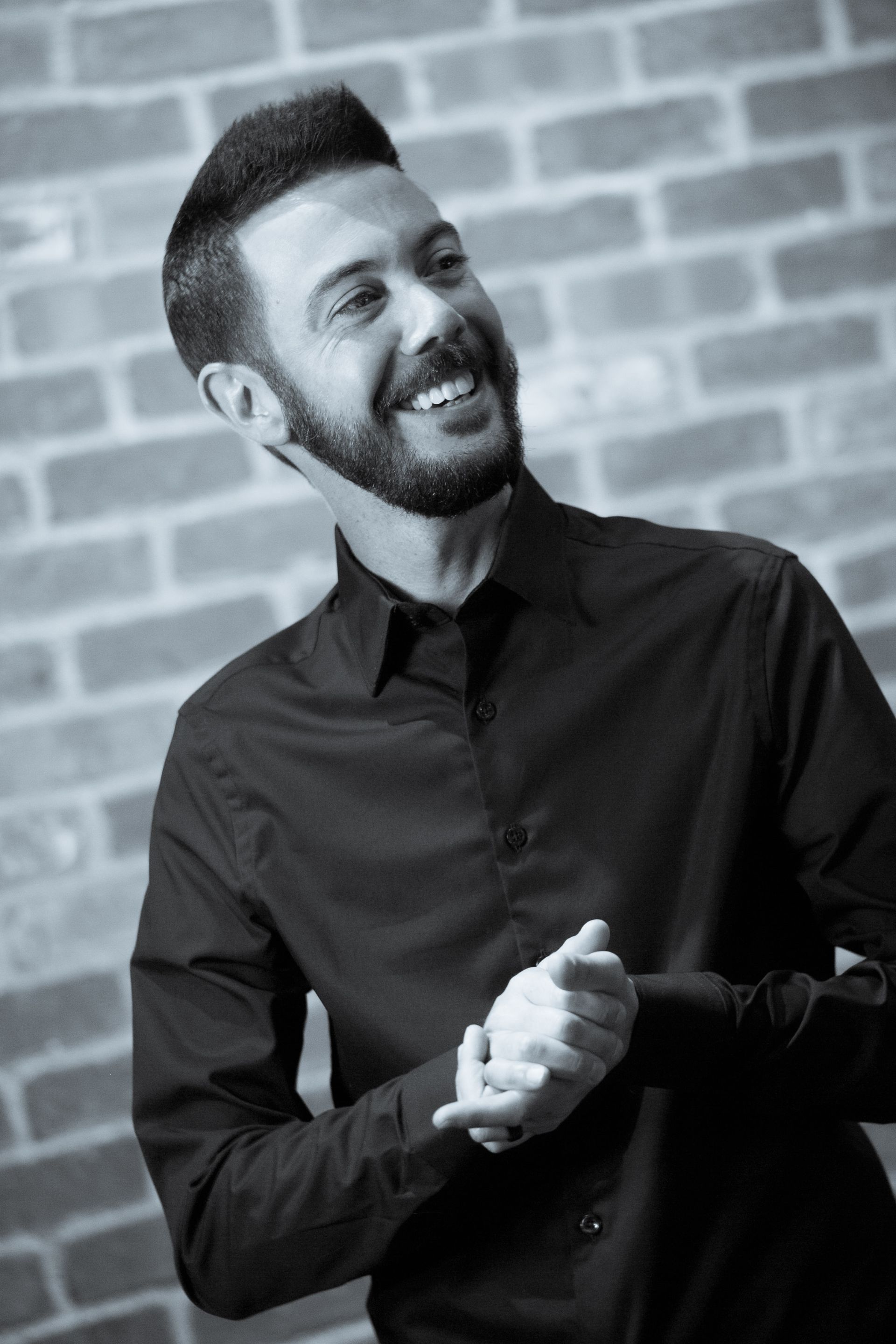 Smiling man in a black shirt standing before a brick wall, hands clasped together — Las Vegas portrait photography