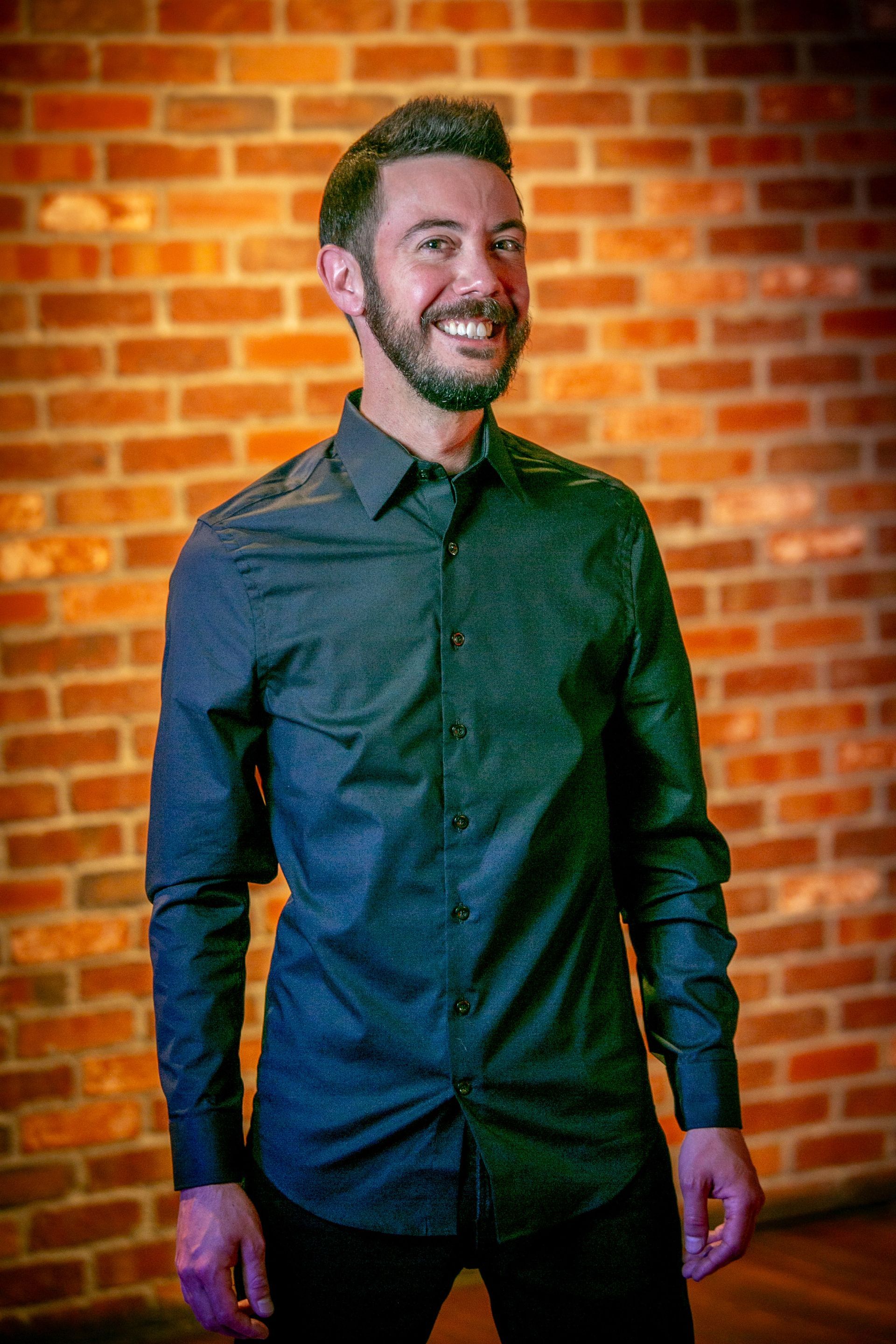 Smiling man in dark button-up shirt standing against a warm brick wall — portrait photography