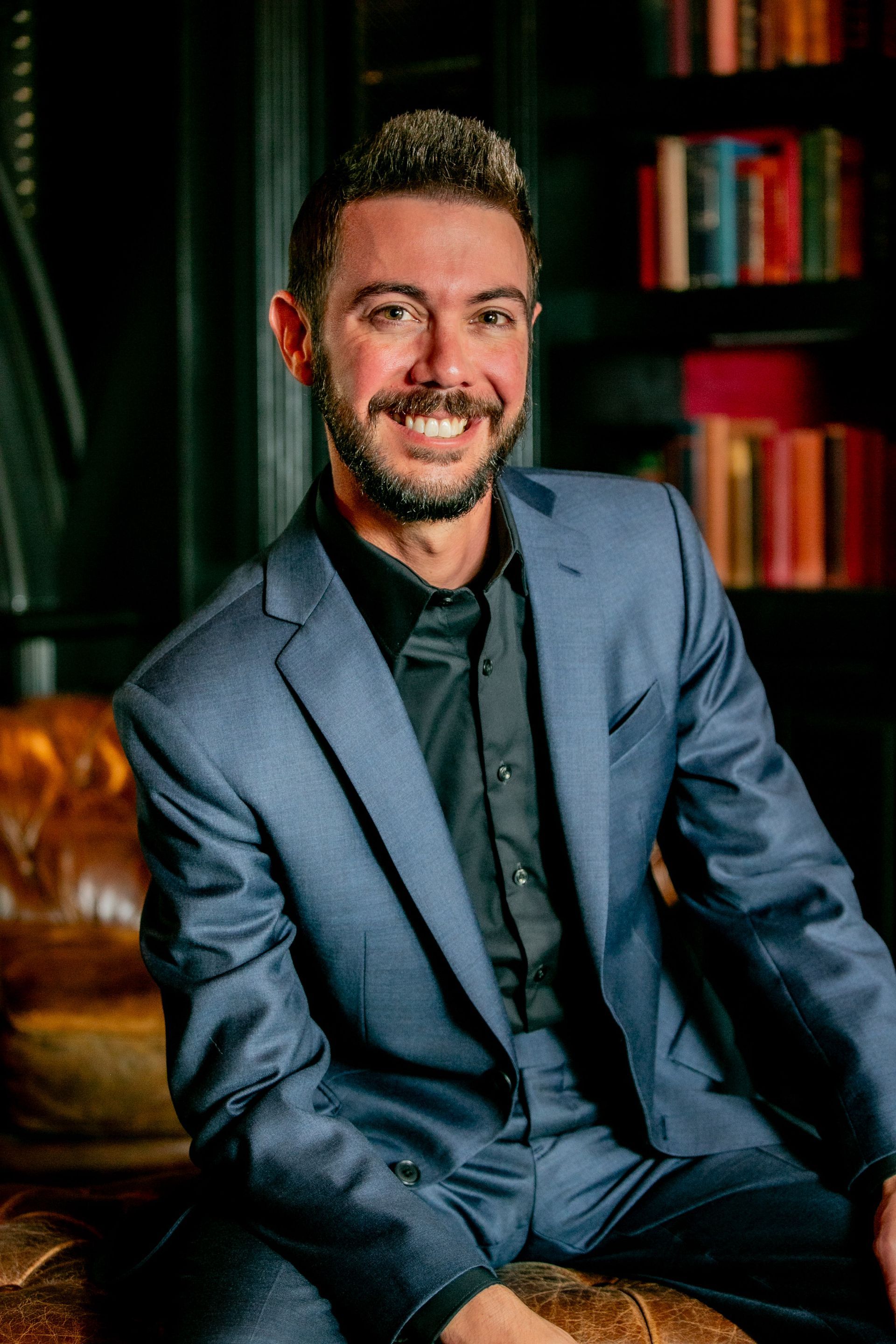 Smiling man in a blue suit seated in a warmly lit room with bookshelves in the background