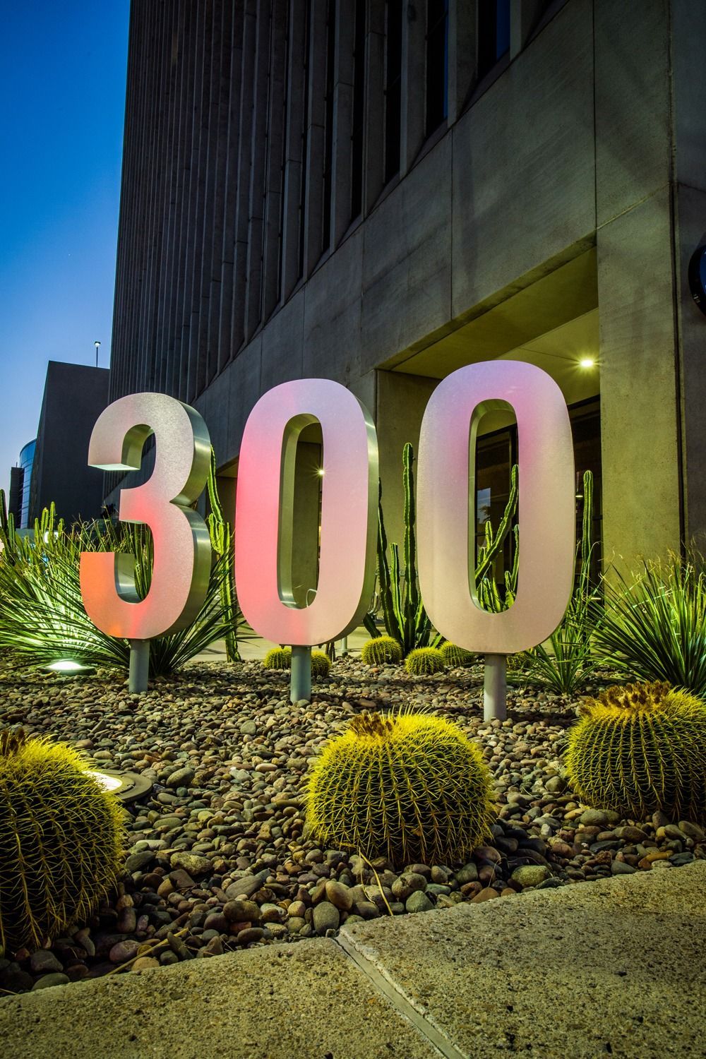 Large pink “300” lawn sign in front of a building at night, surrounded by landscaped greenery — architectural photography