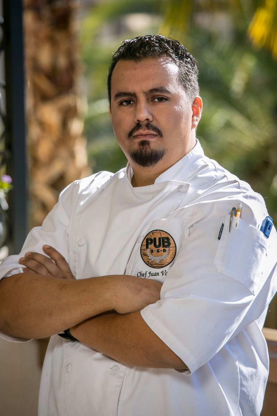 Chef in a white uniform with arms crossed, standing outdoors with a blurred background