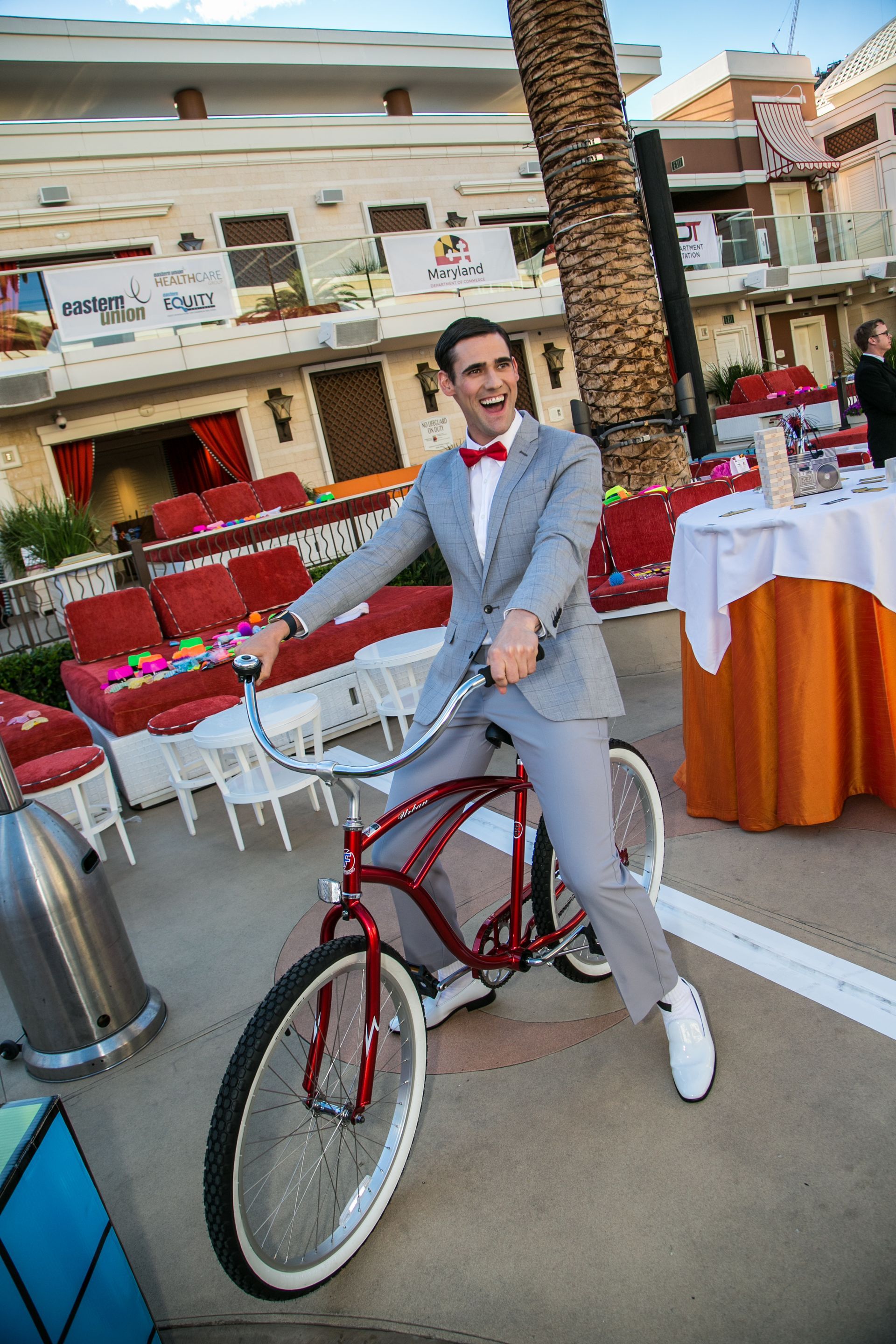 Man posing beside a red bicycle at an outdoor café with white chairs and tables — Las Vegas convention and event photography