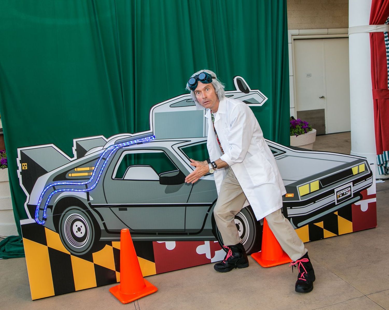 Person posing beside a gray DeLorean model car with orange cones and green backdrop — convention and event photography