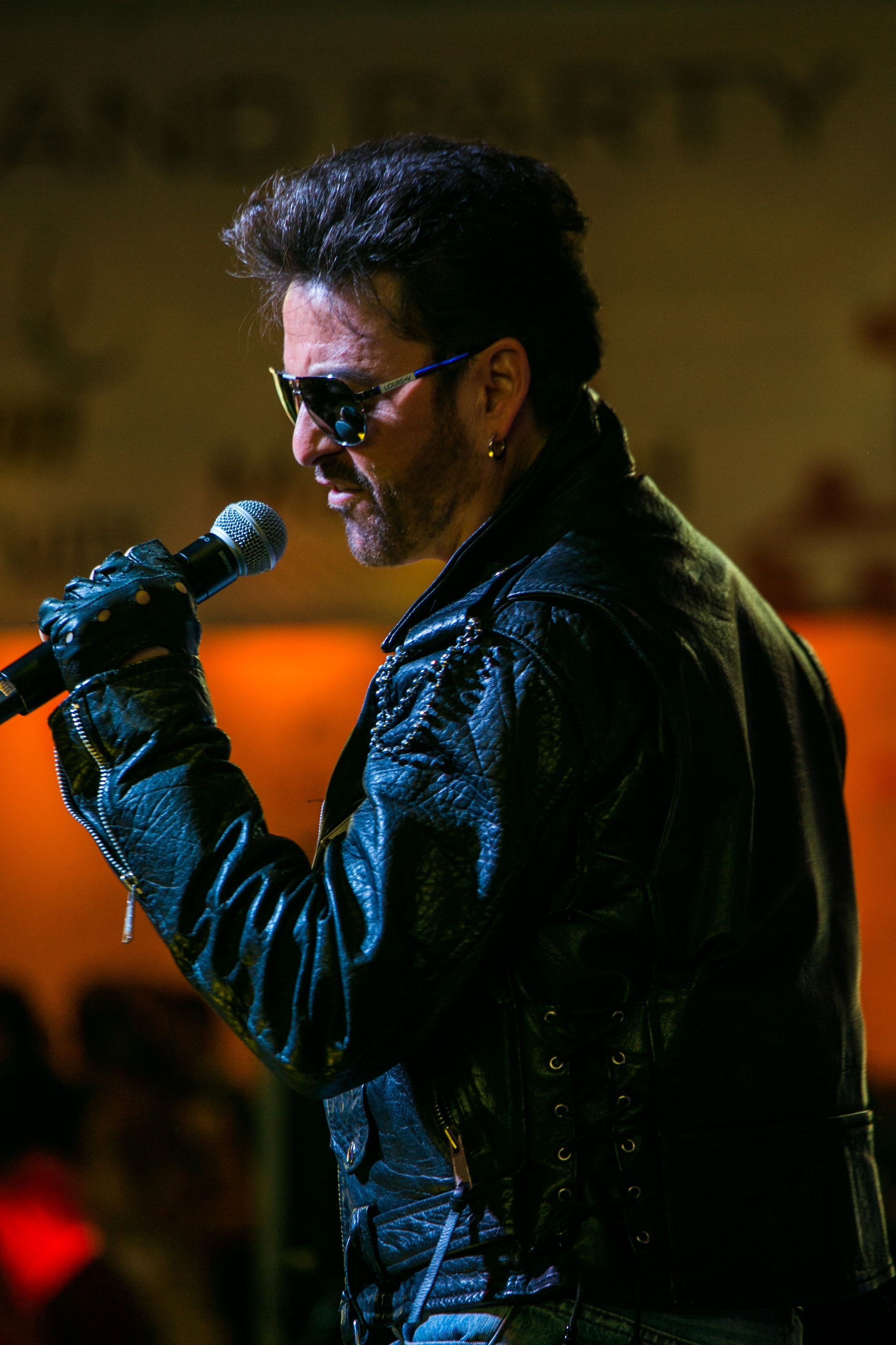 Singer in black leather jacket and sunglasses performs into a microphone onstage — convention and event photography