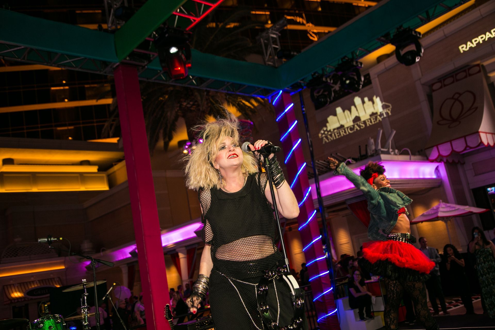 Performer in black outfit and blonde wig on a neon-lit stage, with dancers and colorful club lighting
