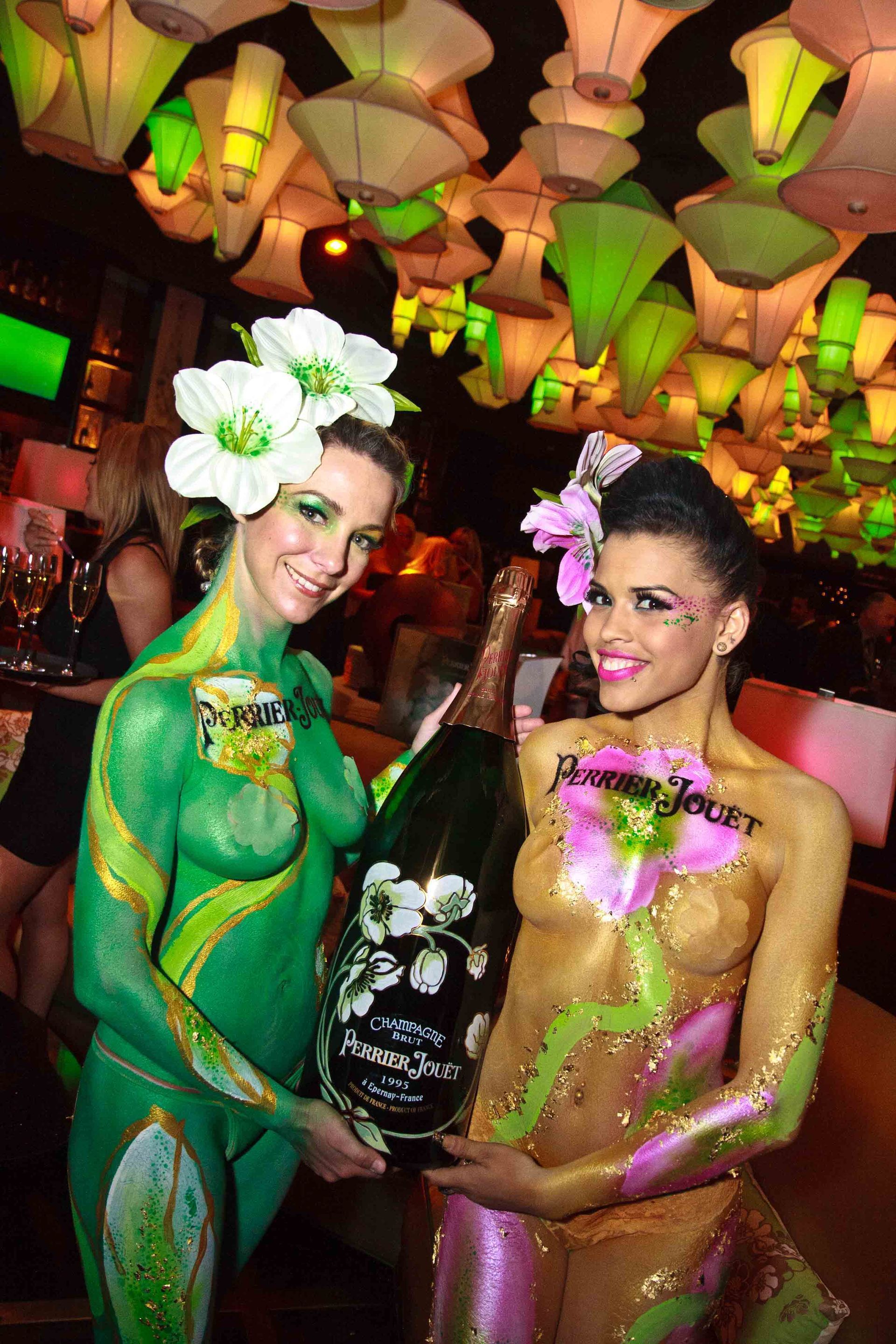 Two women in colorful floral body paint holding a sign, standing under hanging lanterns — convention and event photography
