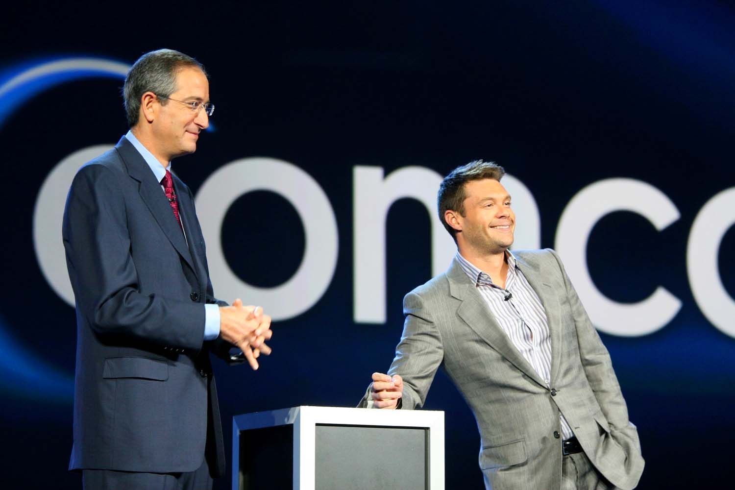 Two men on a stage beside a podium, with a large event logo in the background — convention and event photography