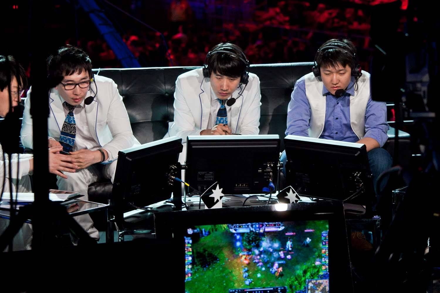 Three esports commentators wearing headsets at a broadcast desk, watching a game on multiple monitors