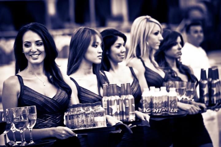 Six women in evening dresses serving drinks at a bar counter, smiling in a dimly lit lounge — Las Vegas convention and event photography