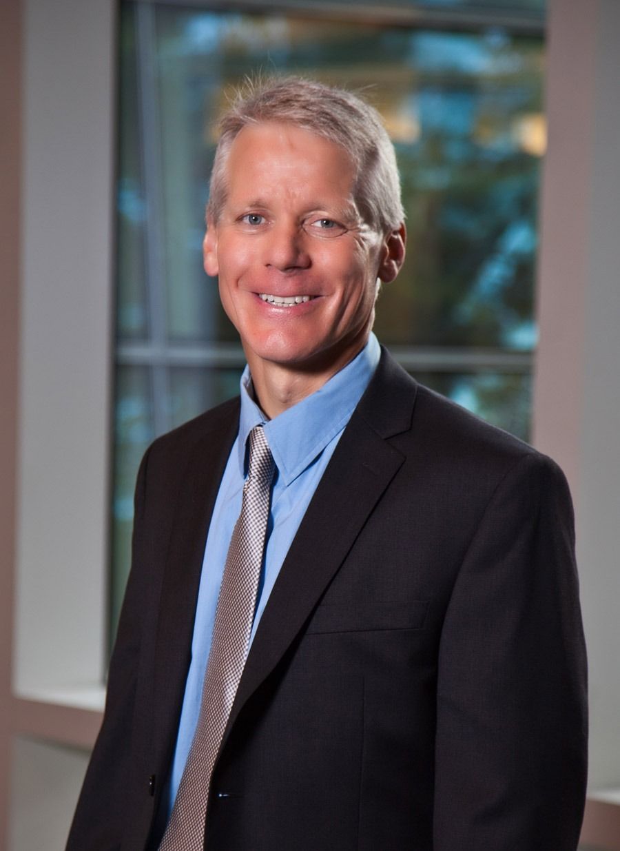 Man in a dark suit and tie standing indoors, smiling in front of a windowed office background — portrait photography