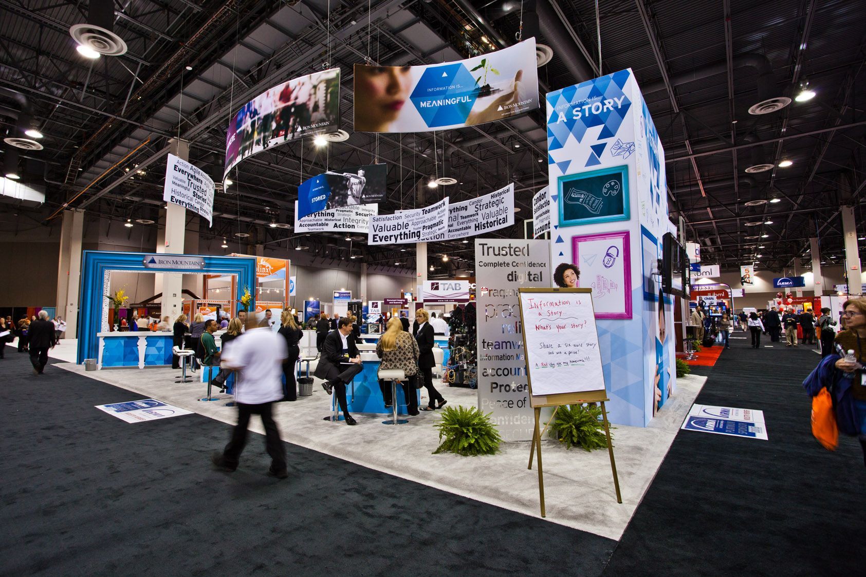 Busy trade show booth with large hanging screens, white display walls — event photography