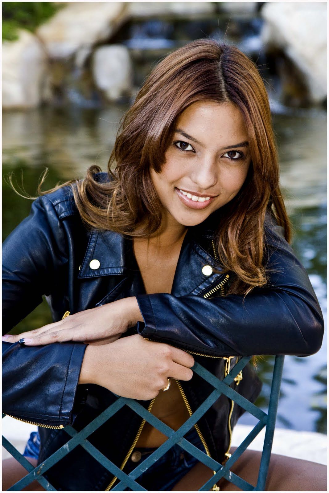 Smiling woman in a black leather jacket leaning on a green fence by water — portrait photography