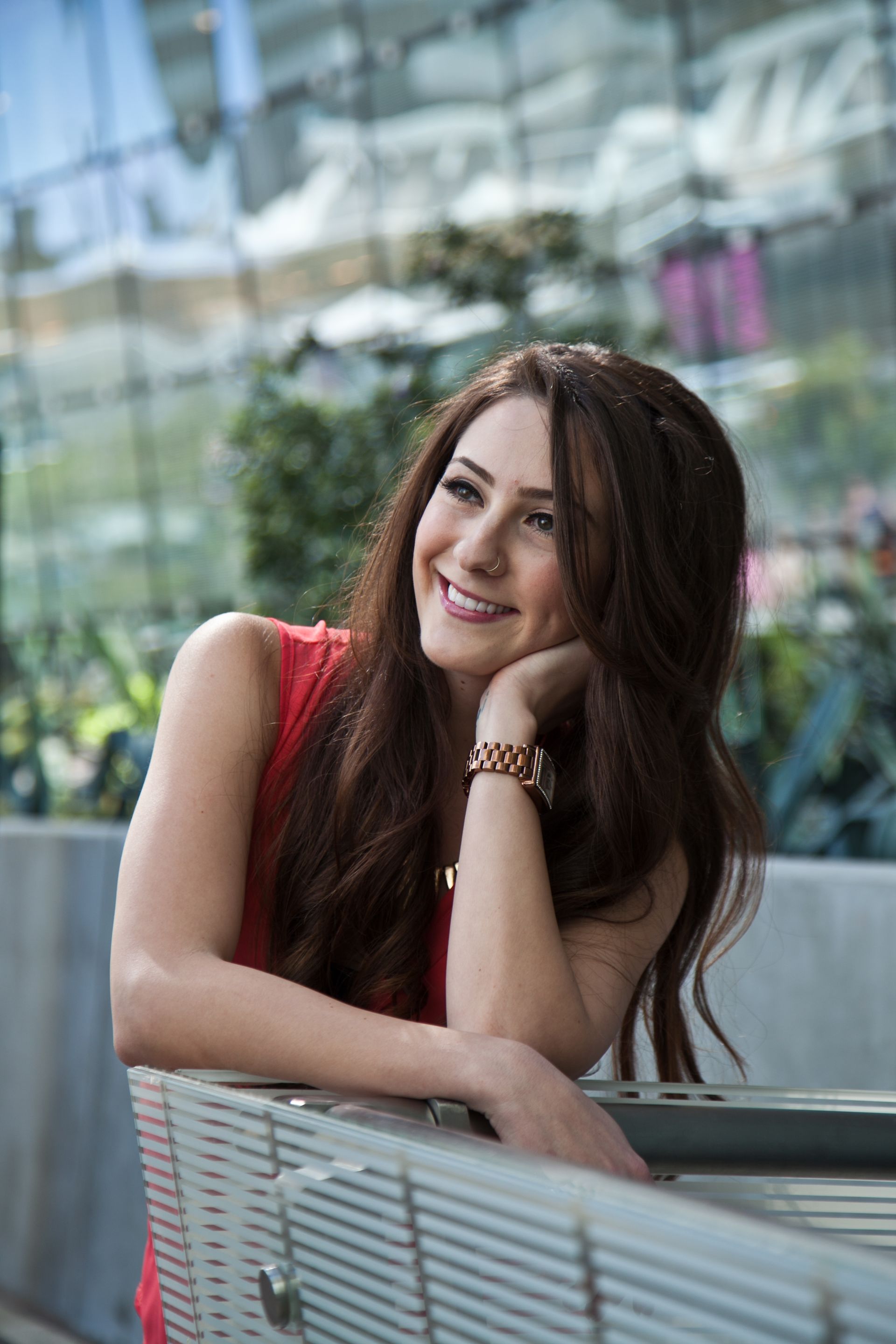 Smiling woman with long brown hair leaning on a bench in a bright glass-walled atrium — portrait photography