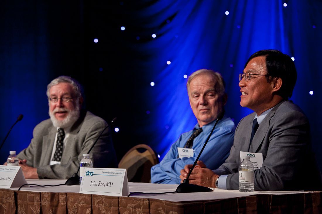 Three men seated at a panel table on a stage with blue lighting, speaking into microphones