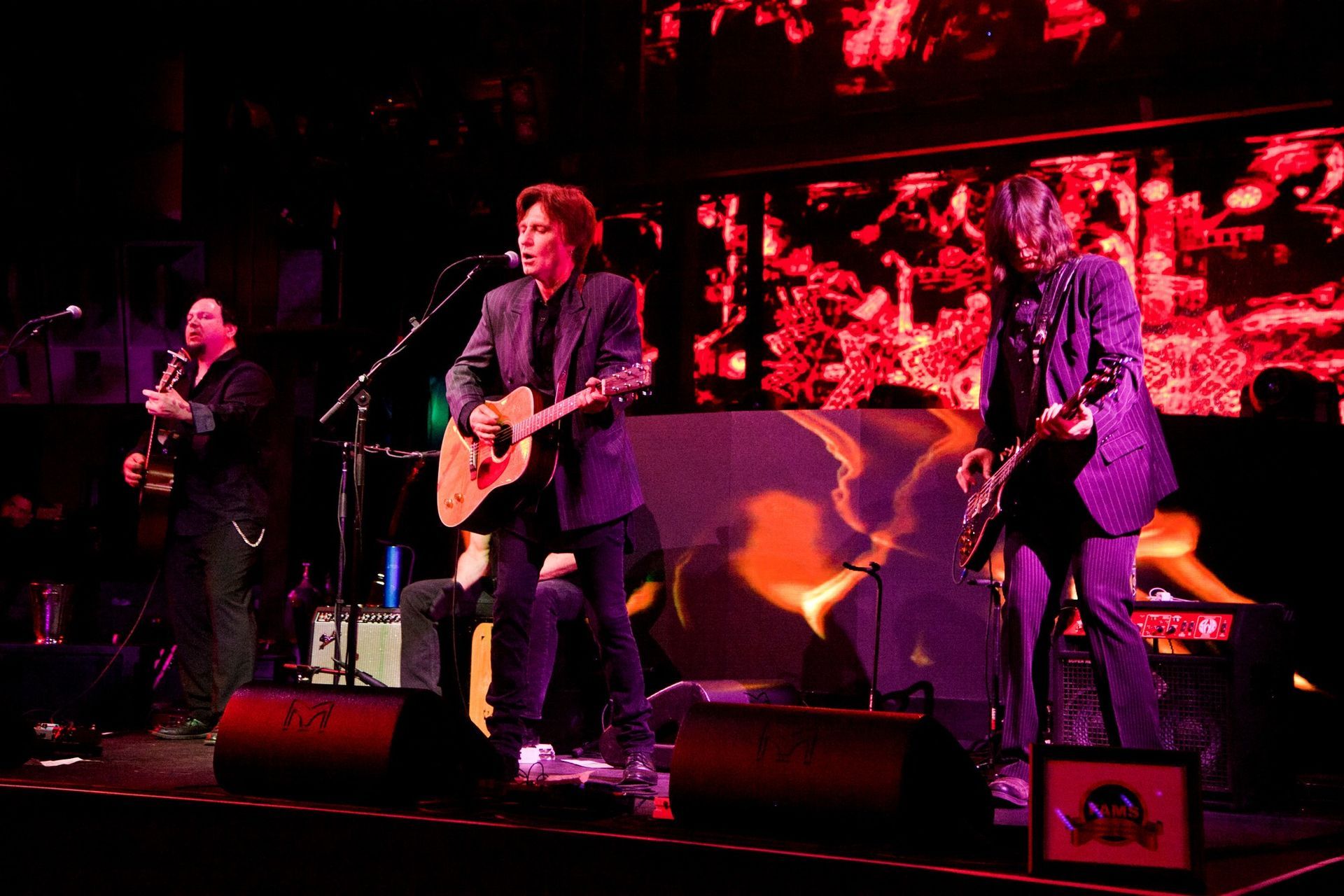 Live rock band performing on stage under red and purple lighting, with guitars and drums visible — professional photography