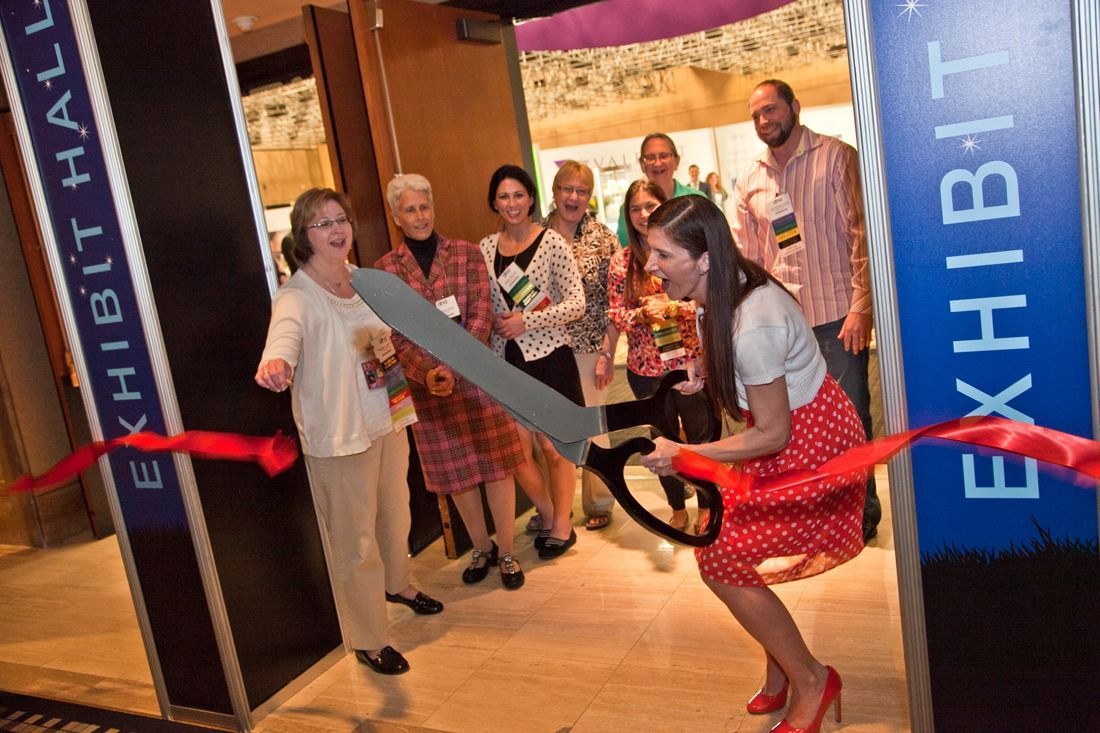 Ribbon-cutting at an exhibit hall entrance, with a group posing behind a woman holding oversized scissors
