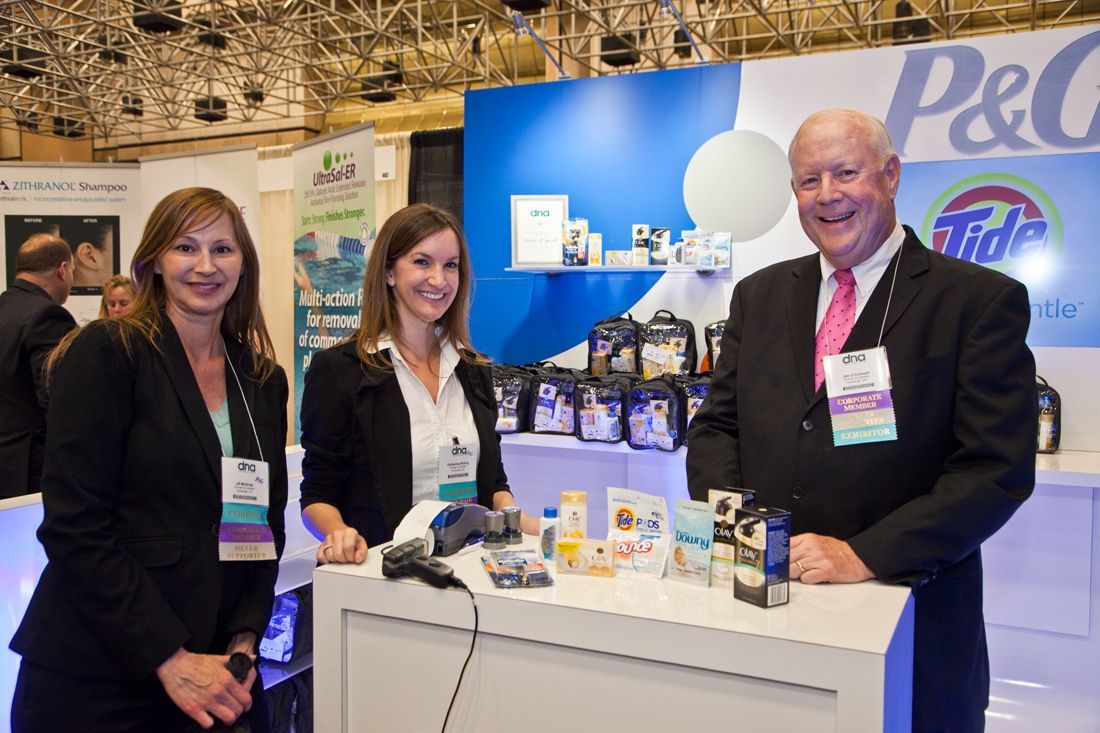 Three people at a trade show booth with product displays and a blue P&G backdrop — convention and event photography