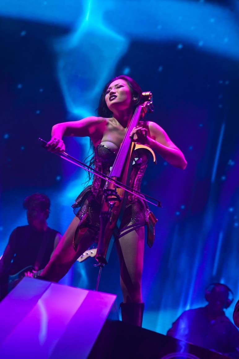 Cellist in a purple-lit stage performance, playing energetically on a cello under blue and pink lights