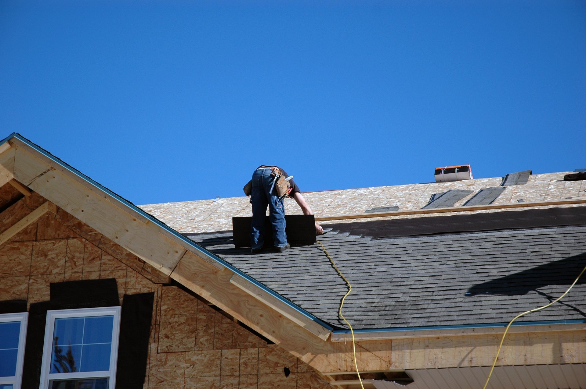 Worker on a roof installing shingles under a clear blue sky.