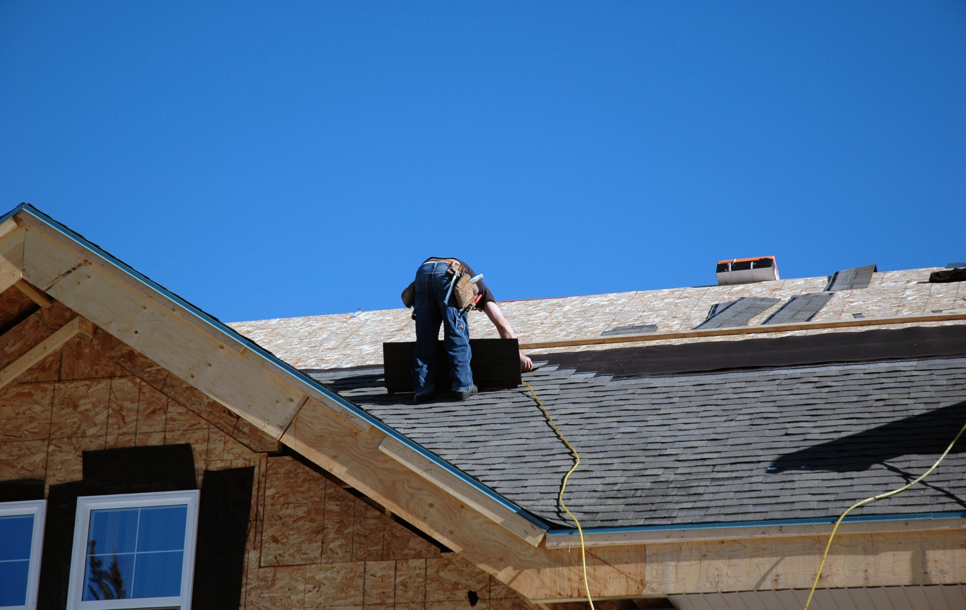 Worker on a shingled roof using a hose against a clear blue sky