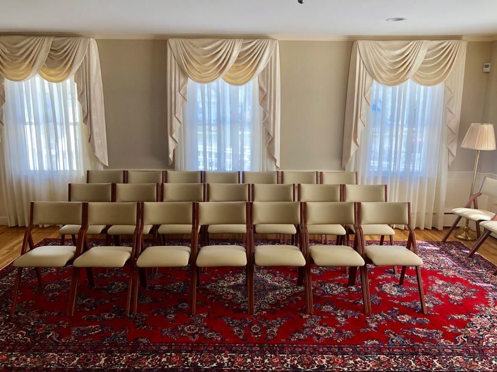 Rows of beige chairs in a formal room with tall windows, cream drapes, and a red patterned carpet.