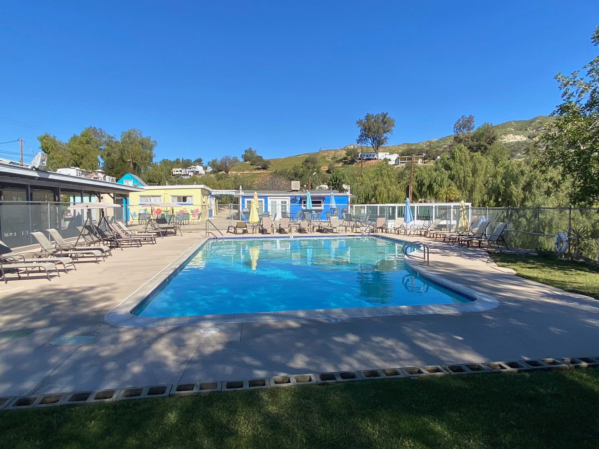 An outdoor swimming pool surrounded by lounge chairs, with small blue and yellow buildings and a grassy hillside behind.