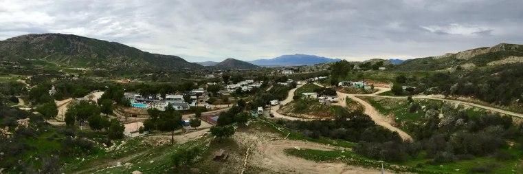 A panoramic view of a small town nestled in a valley among rolling, green, tree-covered hills under a cloudy sky.