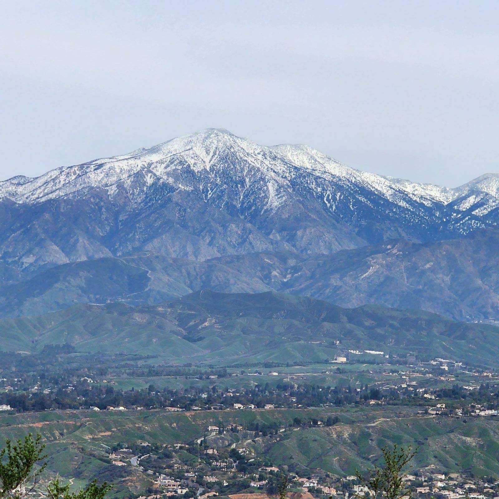 A snow-capped mountain range rises behind a valley containing a sprawling residential neighborhood under a pale sky.