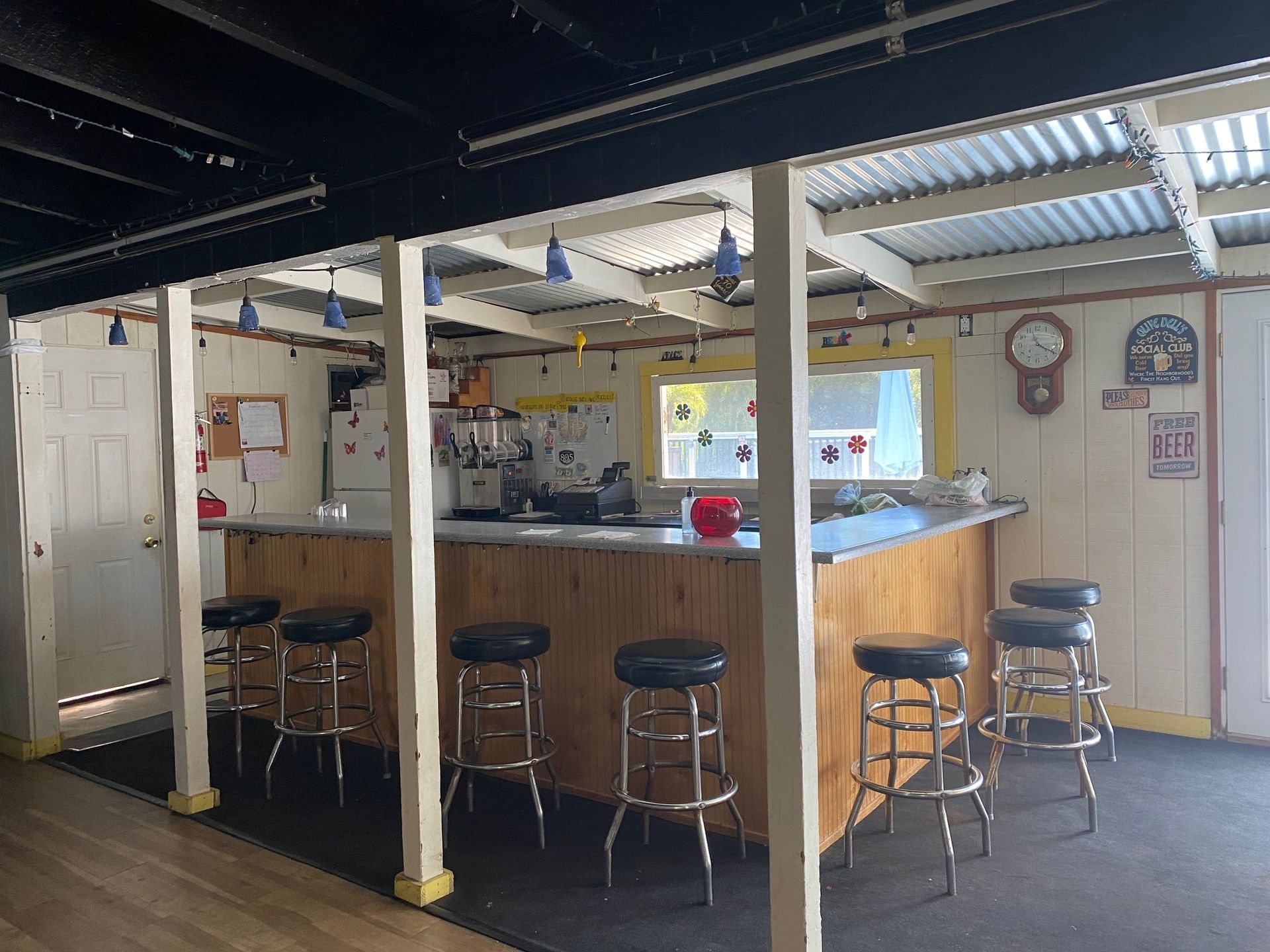 A home bar setup with a wooden counter, five black bar stools, and overhead lighting in a room with a corrugated roof.