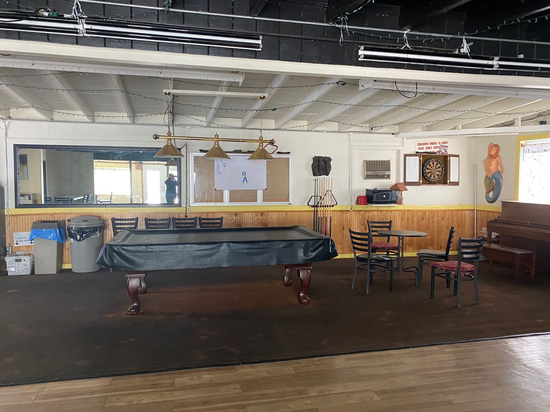 A room with a pool table covered by a dark cloth, a small table with chairs, a dartboard, and a piano.