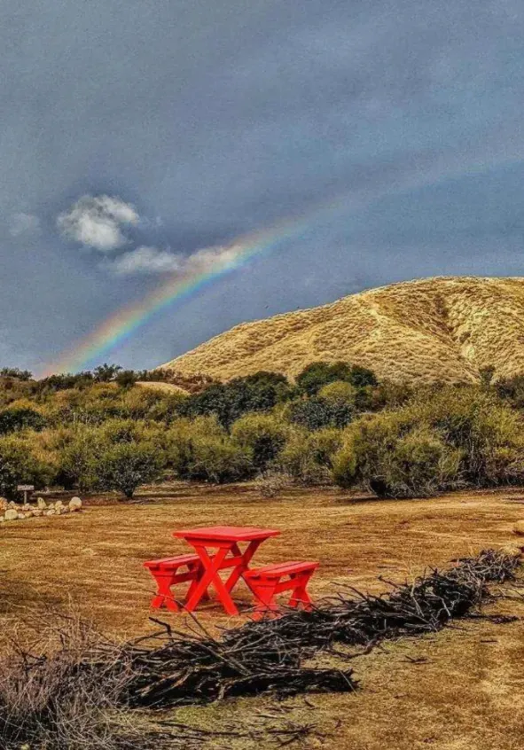 A red picnic table sits in a dry, open field before a hill, with a faint rainbow arching across a cloudy, stormy sky.