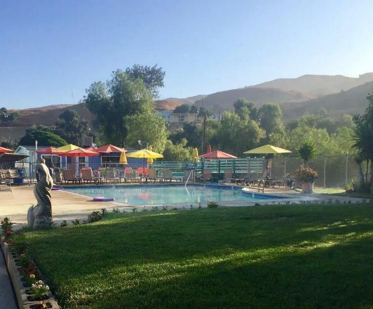 A swimming pool surrounded by yellow and orange umbrellas, green grass, and trees with desert mountains in the background.