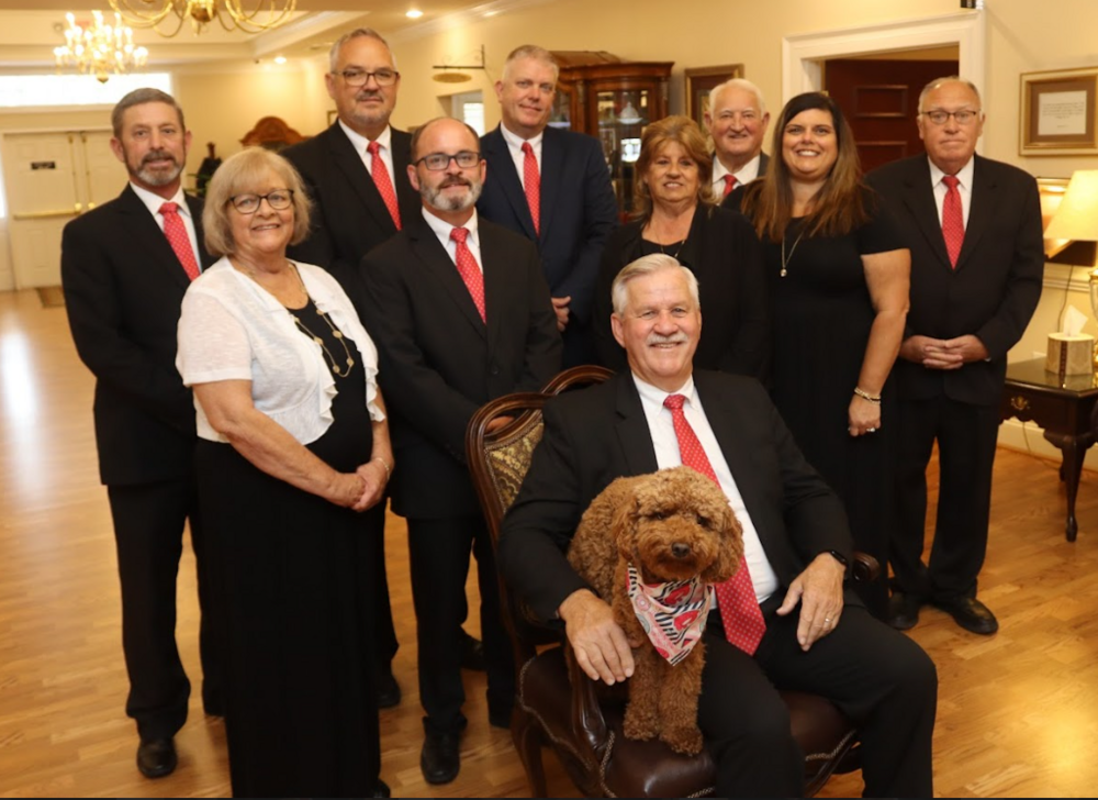 Group portrait of ten people in formal attire, with a dog seated in front, in a wood-paneled room